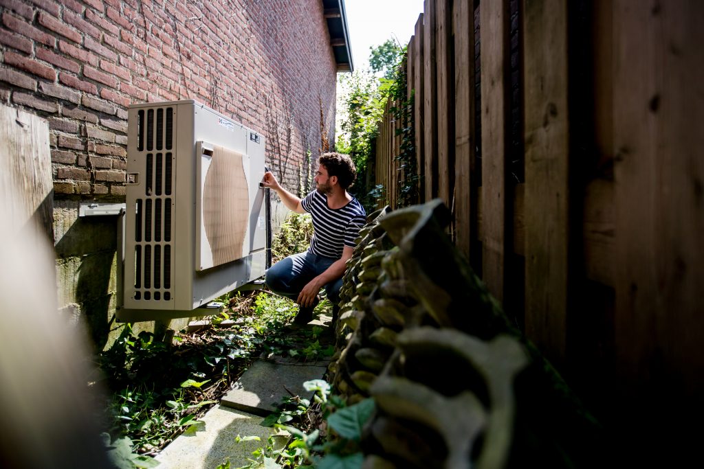 Een man zit gehurkt voor een warmtepomp die p de buitengevel van zijn huis is bevestigd.