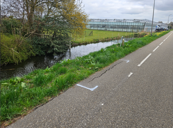 een weg met bermen met groen gras, kassen en bomen er langs waarbij een stukje asfalt kapot is en gerepareerd moet worden