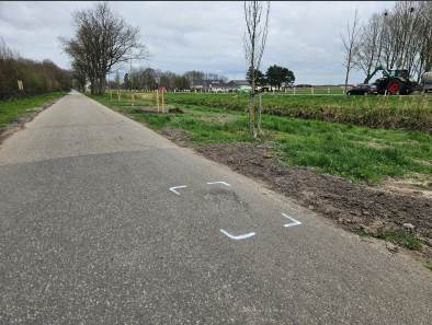 een weg met bermen met groen gras en bomen er langs waarbij een stukje asfalt kapot is en gerepareerd moet worden