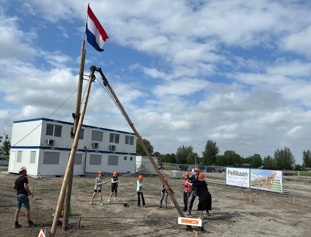 Mensen trekken aan touwen die aan palen zitten om een vlag te hijsen. Zo slaan ze de eerste paal voor een nieuw gebouw