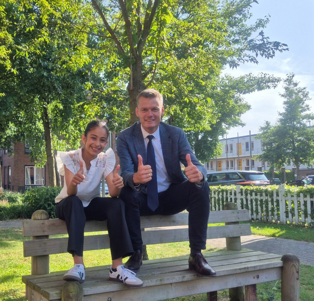 Kinderburgemeester Amira en burgemeester Nieuwenhuis zitten op een houten bankje en houden duimen omhoog