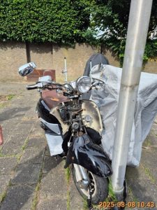 Een zwarte scooter met bruin zadel zonder kenteken, Floriserf vlakbij nummer 2 op de parkeerplaats.