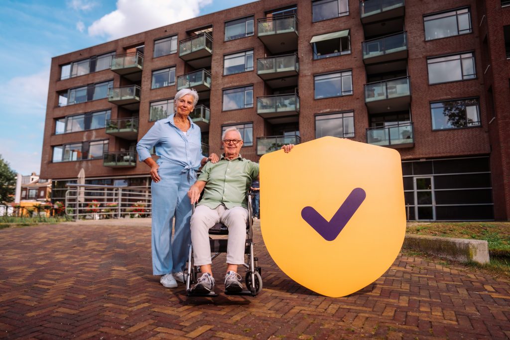 man in rolstoel met flat op de achtergrond en vrouw staat naast rolstoel