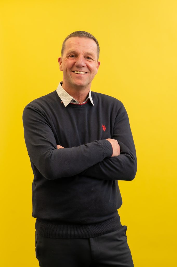 Man staat glimlachend met armen over elkaar voor een gele achtergrond.