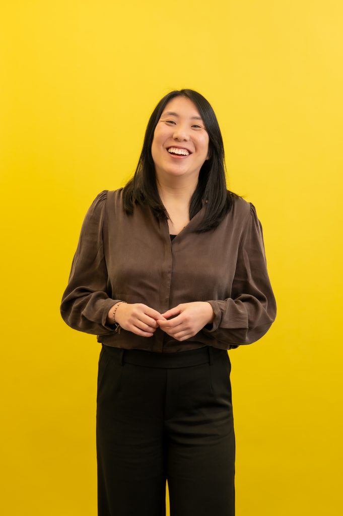 Vrouw staat glimlachend met gevouwen handen voor een gele achtergrond.